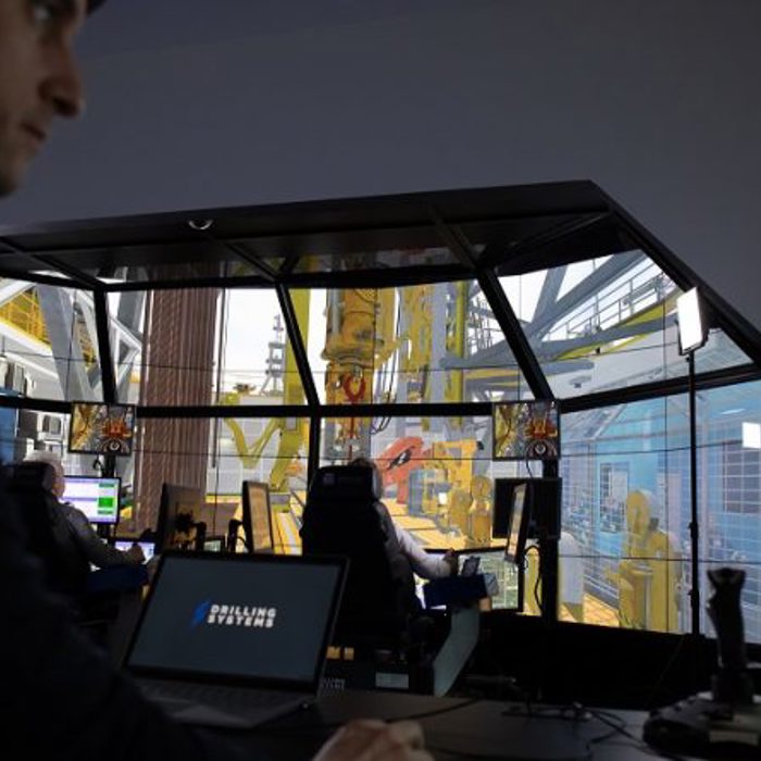 Person operating drilling simulation software in a control room equipped with multiple computer screens and a drilling rig simulator.