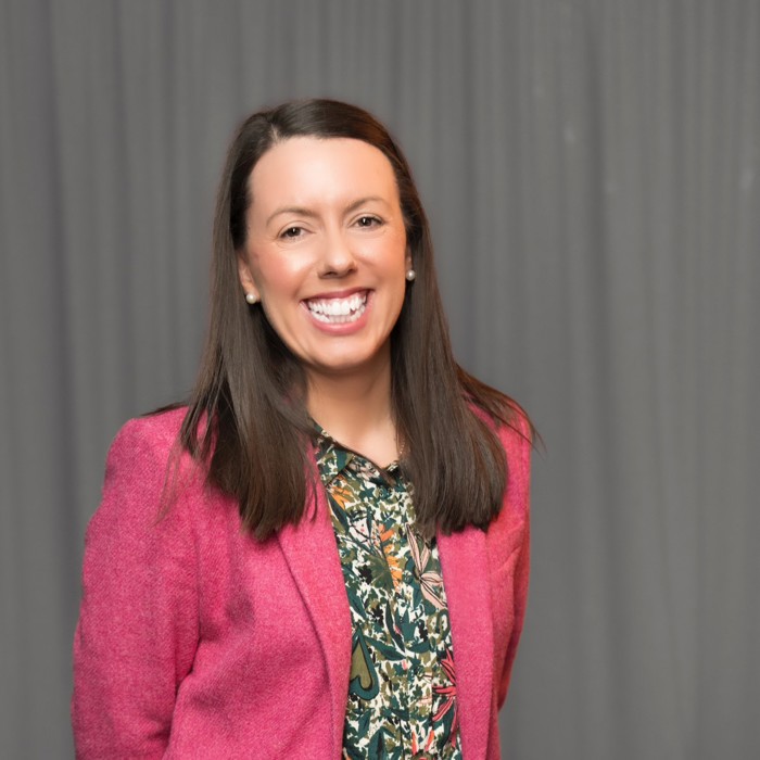 A woman with long dark hair is smiling, wearing a pink blazer over a patterned blouse, and standing in front of a gray curtain.