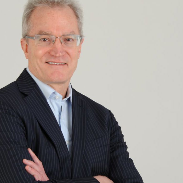 A man with short gray hair, wearing glasses, a light blue shirt, and a pin-striped dark suit, stands against a plain background with arms crossed, smiling at the camera.