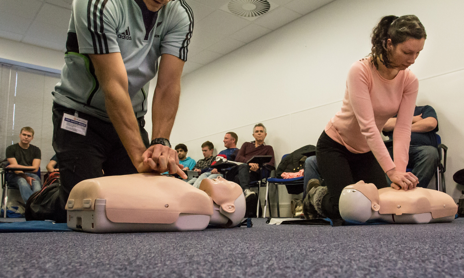 In a training session, a group of people in a classroom setting practice CPR on mannequins. Two individuals in the foreground are kneeling and performing chest compressions with precision and focus.
