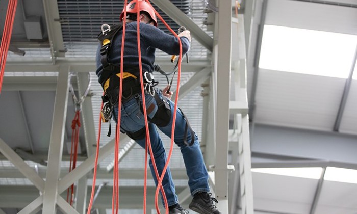 Une personne portant un équipement de sécurité et un casque rouge est suspendue à des cordes, grimpant sur une structure métallique à l'intérieur d'une installation industrielle.