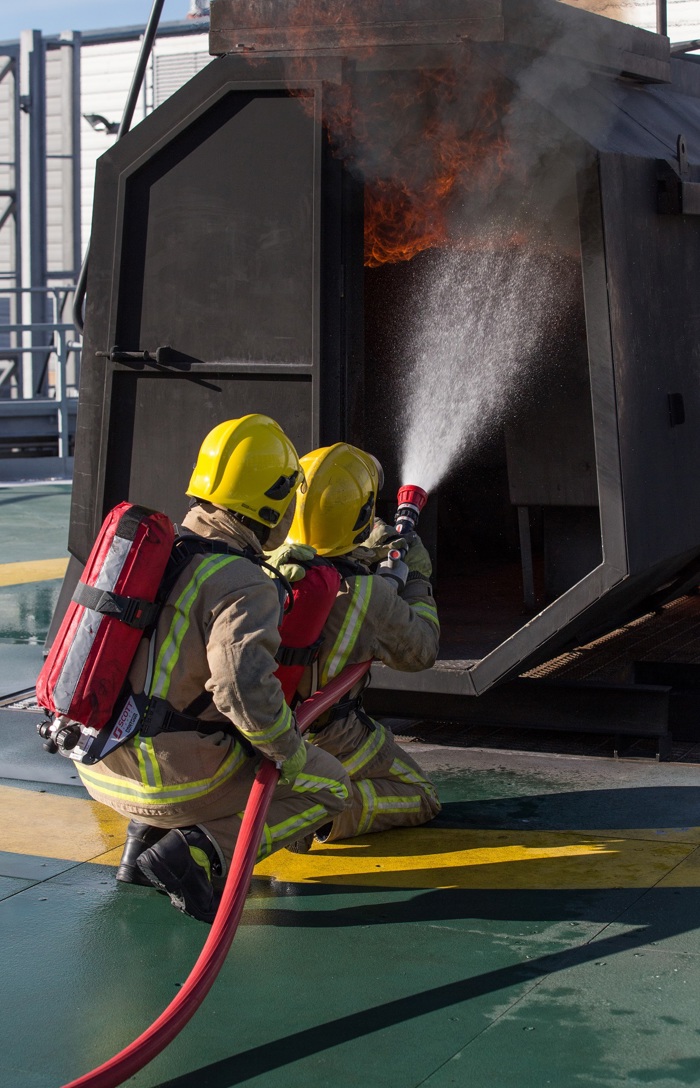 Bomberos con cascos amarillos y equipo de protección rocían agua en una estructura en llamas durante un ejercicio de entrenamiento.