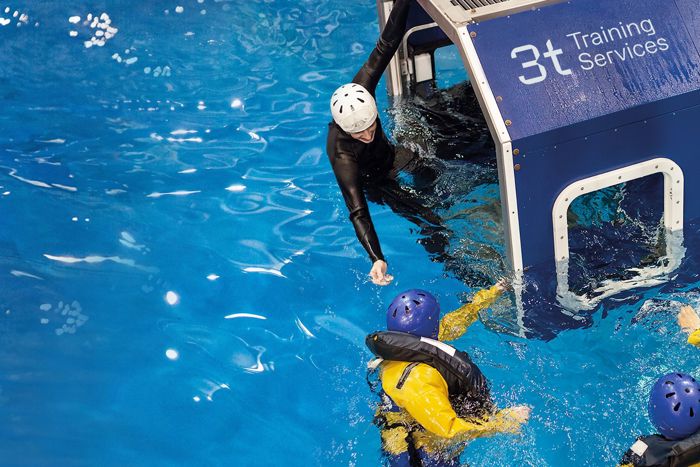 Two individuals in the water perform a safety exercise with one person reaching out from a cabin labeled "3t Training Services" while another person inside wears a helmet.