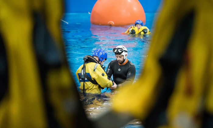 Deux personnes en tenue d'activité aquatique sont vues au centre d'une piscine, entourées d'autres personnes en tenue jaune, avec une grande bouée orange à l'arrière-plan.