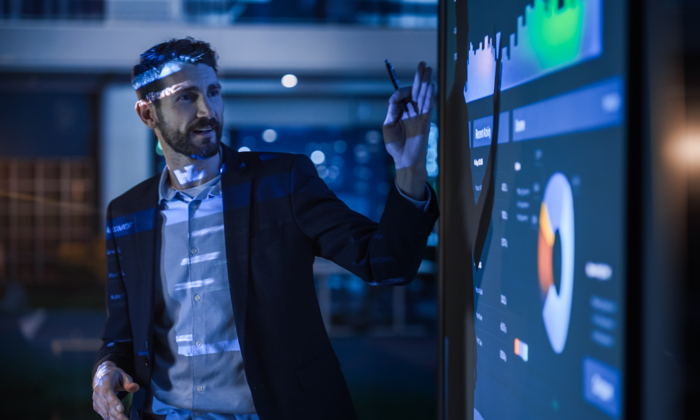 Um homem de fato de trabalho aponta para um grande ecrã interativo que apresenta gráficos e quadros num ambiente de escritório moderno.