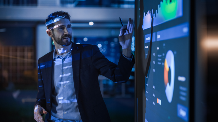 Um homem de fato de trabalho aponta para um grande ecrã interativo que apresenta gráficos e quadros num ambiente de escritório moderno.