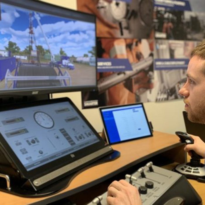 A person operates a machine simulator with multiple screens displaying controls and a simulated outdoor drilling scene.