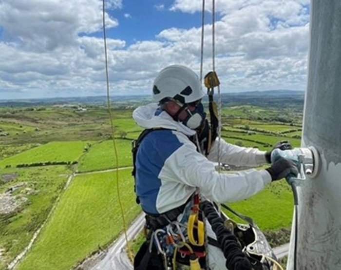 Uma pessoa com equipamento de segurança e capacete trabalha numa estrutura no alto de uma paisagem rural com campos verdes sob um céu parcialmente nublado.