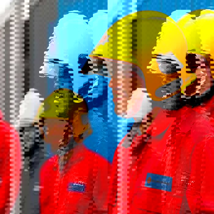 A group of four people wearing yellow helmets and red uniforms are standing outside beside blue structures.