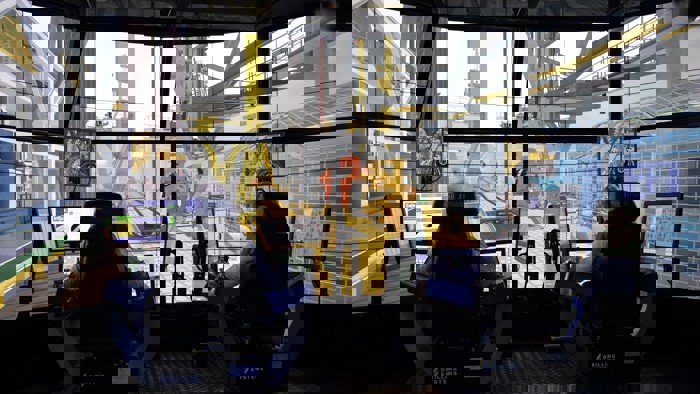 Two people sit at control stations with multiple monitors, operating a high-tech drilling simulator. Large screens simulate an offshore drilling environment around them.