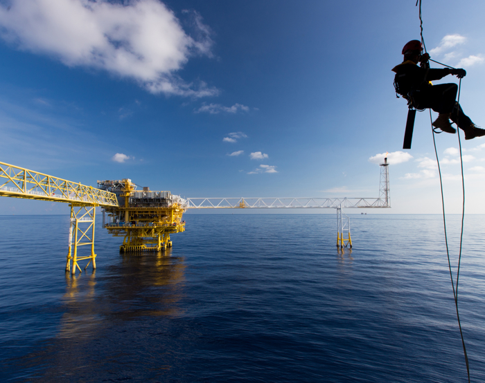 A worker, undergoing training, is suspended by a safety rope above the ocean, with an offshore oil platform in the background under a blue sky with scattered clouds.