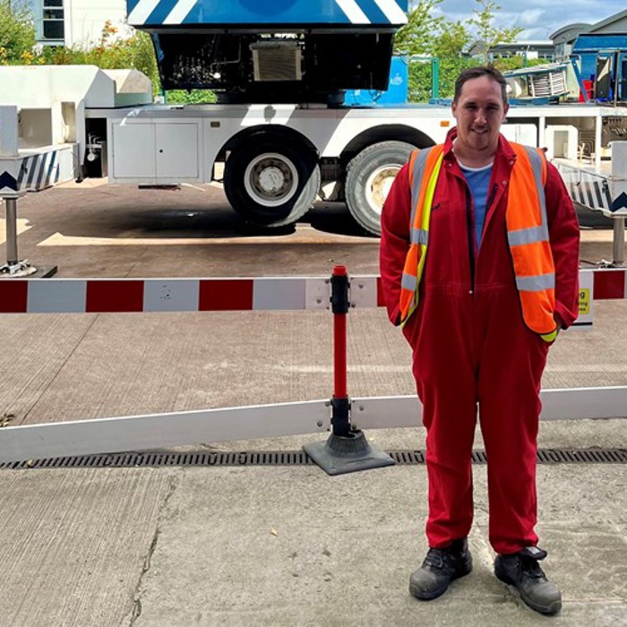 Person in a red jumpsuit and reflective safety vest stands in front of a barrier, with a large industrial vehicle in the background.