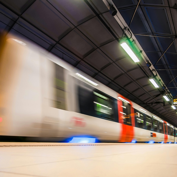 A fast-moving train passes through an underground station, with overhead signs visible. The image captures the motion blur of the train.