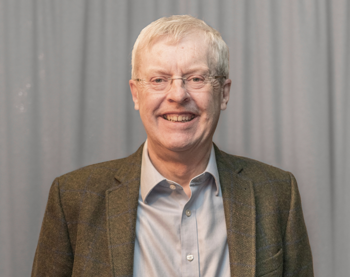 Um homem mais velho, de óculos e casaco cinzento, sorri com confiança, possivelmente reflectindo sobre anos de treino, em frente a uma cortina cinzenta.