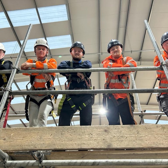 scaffolding apprentices in hard hats and harnesses looking down from scaffolding platform