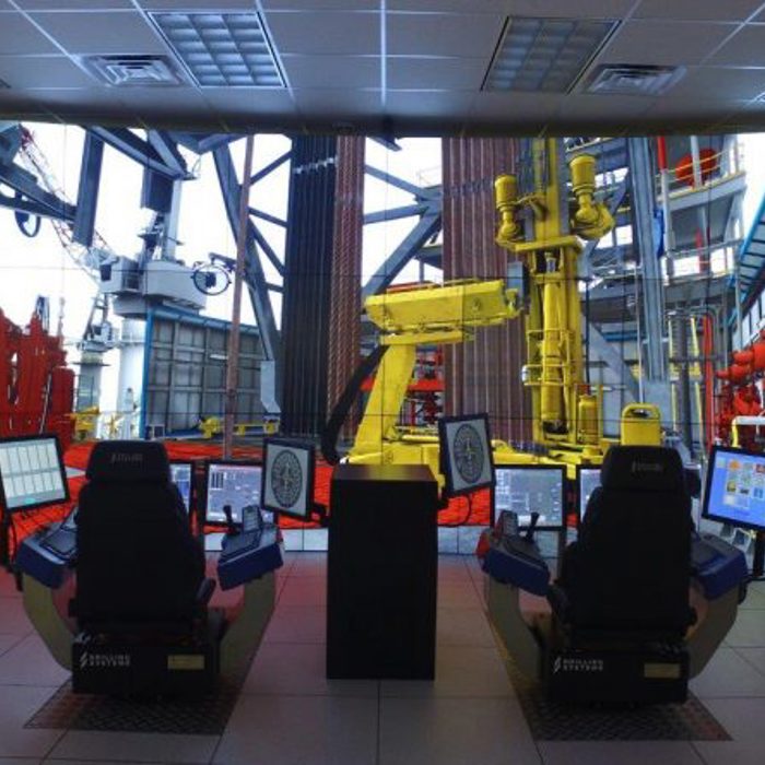 Two control stations with screens and joysticks face a large display showing an industrial machinery environment, likely simulating remote operations. Multiple monitors are mounted on the surrounding walls.