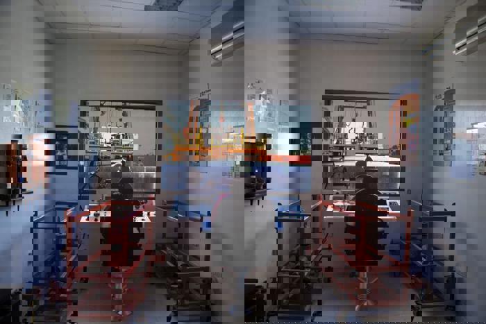 Interior de una sala de control con una persona sentada ante una consola, vigilando una grúa que eleva un contenedor en el exterior. La sala está flanqueada por bastidores rojos y en las paredes hay carteles informativos.