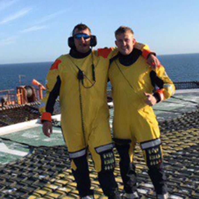 Two individuals in yellow protective suits stand side by side on a helipad offshore, with an expanse of the ocean and clear sky in the background.