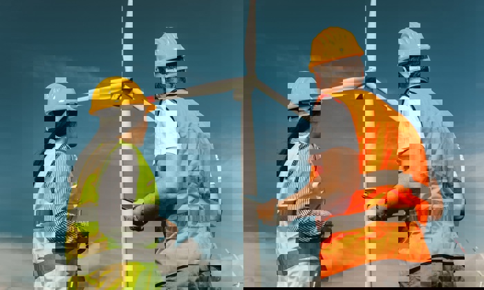 Two workers wearing hard hats and reflective vests stand near a large wind turbine, observing and possibly inspecting it under a clear blue sky.