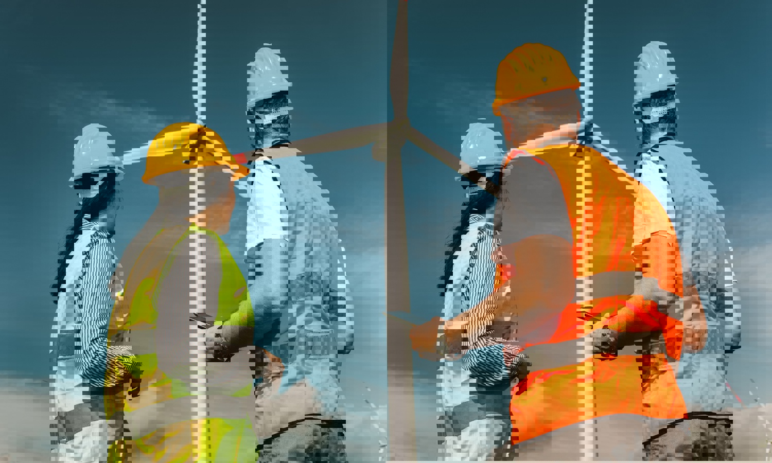 Dois trabalhadores com capacetes e coletes reflectores estão perto de uma grande turbina eólica, observando-a e possivelmente inspeccionando-a sob um céu azul claro.