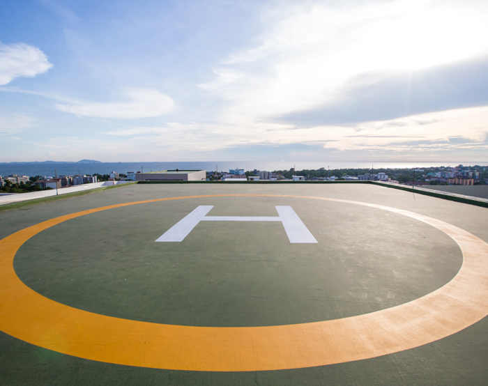 A rooftop helipad with a white "H" in the center is shown under a partly cloudy sky with a distant view of buildings and hills.