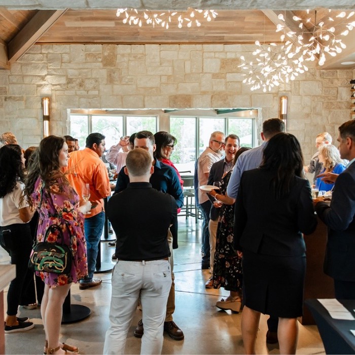 A group of people socializing in a spacious room with large windows, stone walls, and modern lighting, with wine bottles displayed on a rack in the background.
