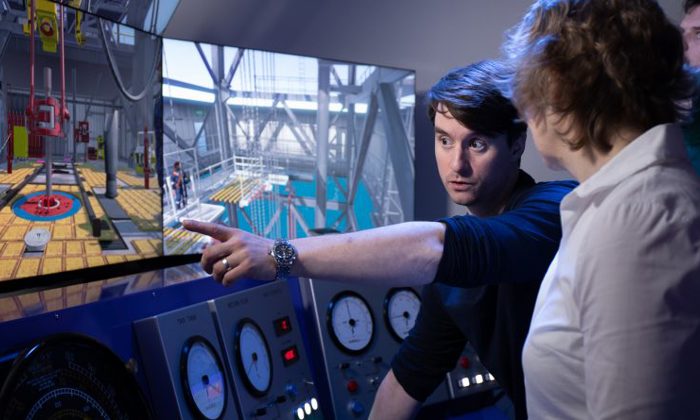 Uma pessoa aponta para um ecrã que apresenta controlos industriais e simulações de máquinas enquanto explica a outra pessoa em frente a painéis de controlo com medidores analógicos.