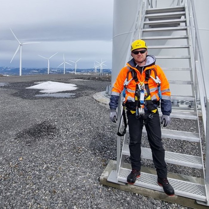 A person in safety gear stands at the base of a wind turbine, with multiple wind turbines visible in the background on a gravel-covered landscape.