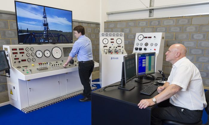 Two men operate an oil and gas drilling simulator in a control room, with one seated at a computer desk and the other standing at control panels. A large screen displays a drilling rig.