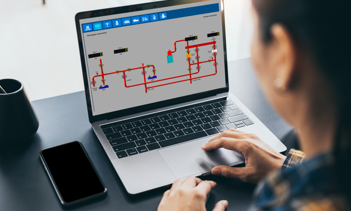 Uma pessoa utiliza um computador portátil que apresenta um diagrama esquemático de um sistema técnico com tubagens e instrumentos. Um smartphone e uma chávena estão sobre a secretária.
