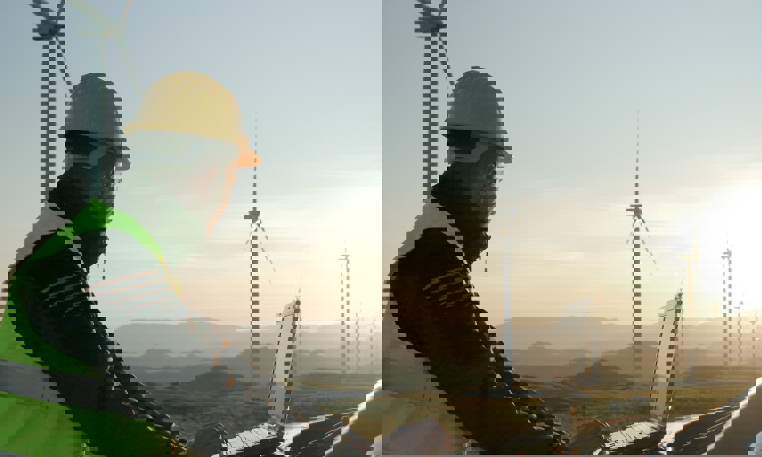 شخص يرتدي قبعة صلبة وسترة أمان ويستخدم جهاز كمبيوتر محمول في الهواء الطلق بالقرب من توربينات الرياح، مع مناظر طبيعية خلابة للتلال في الخلفية أثناء غروب الشمس.