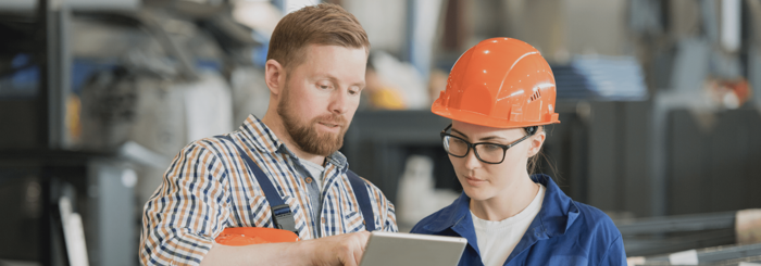 Two workers in a factory setting. One, in plaid shirt, is pointing at a tablet screen, while the other, in a blue uniform and orange hardhat, listens attentively.