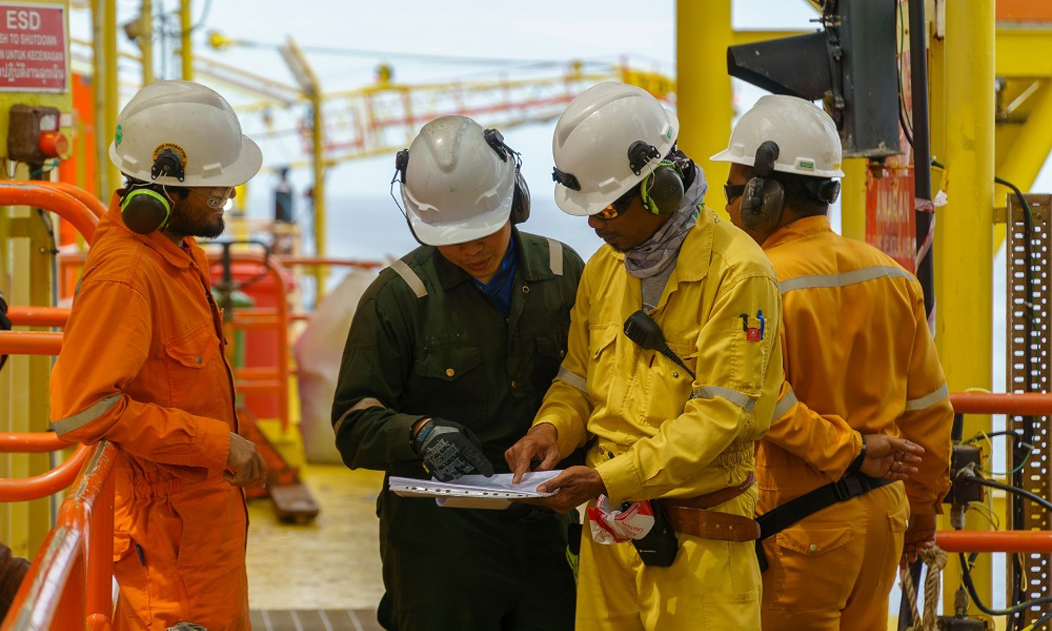 Quatro trabalhadores com equipamento de segurança analisam um documento numa plataforma petrolífera offshore. Dois de fato-macaco cor de laranja, um de preto e um de amarelo discutem o conteúdo do documento enquanto estão junto a grades cor de laranja.