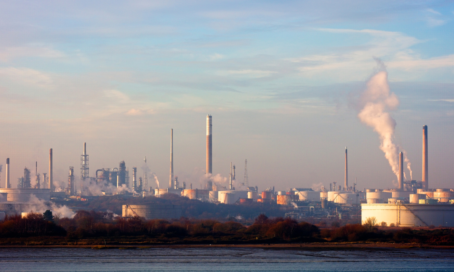 A paisagem de uma refinaria industrial estende-se sob um céu azul com nuvens dispersas, com as suas chaminés a deitar fumo como sentinelas em formação, sincronizadas e firmes.