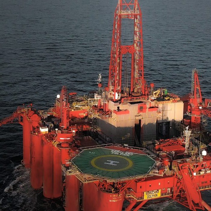 A red offshore oil drilling platform is positioned in open water, displaying a helipad and various equipment on its deck.