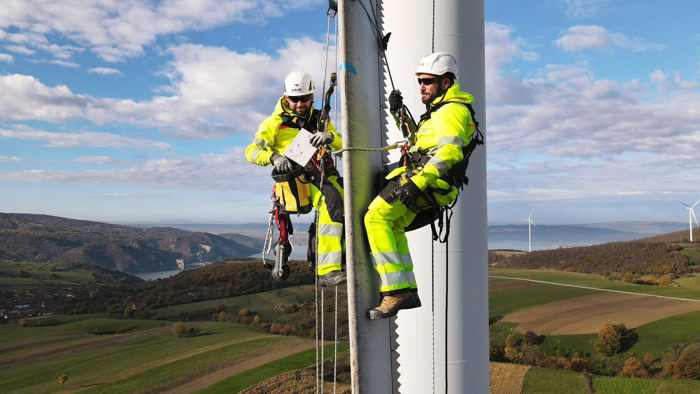 Dois trabalhadores com equipamento de segurança, demonstrando a sua formação, inspeccionam uma turbina eólica enquanto estão presos por cordas, com uma paisagem cénica de colinas e mais turbinas ao fundo.
