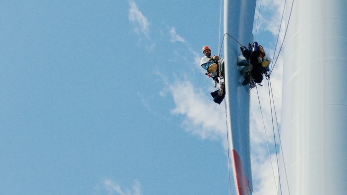 Two workers in safety gear, demonstrating their expert training, are suspended by ropes beside a wind turbine against a blue sky.