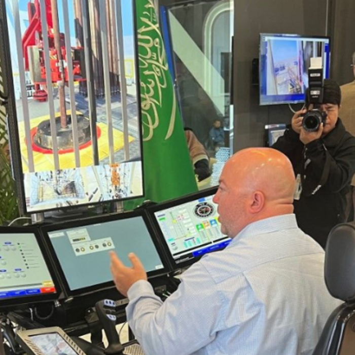 Three men are engaged in conversation near a control panel with multiple screens. One man, wearing a headscarf, faces a seated man who gestures toward the screens. Another man stands nearby.