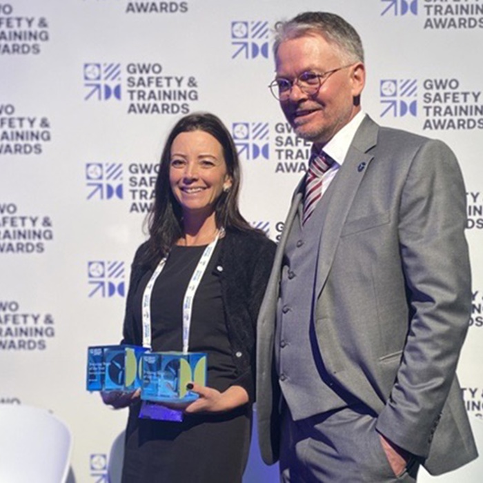 Two people stand in front of a backdrop with "GWO Safety & Training Awards" logos. The woman on the left holds two awards, while the man on the right is dressed in a suit and tie.