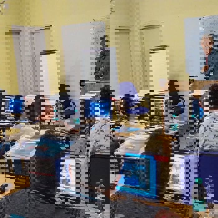 A man stands at a podium, speaking to a classroom of seated adults. The room has computer stations, a whiteboard with notes, and a sign reading "CLASS ROOM AREA.