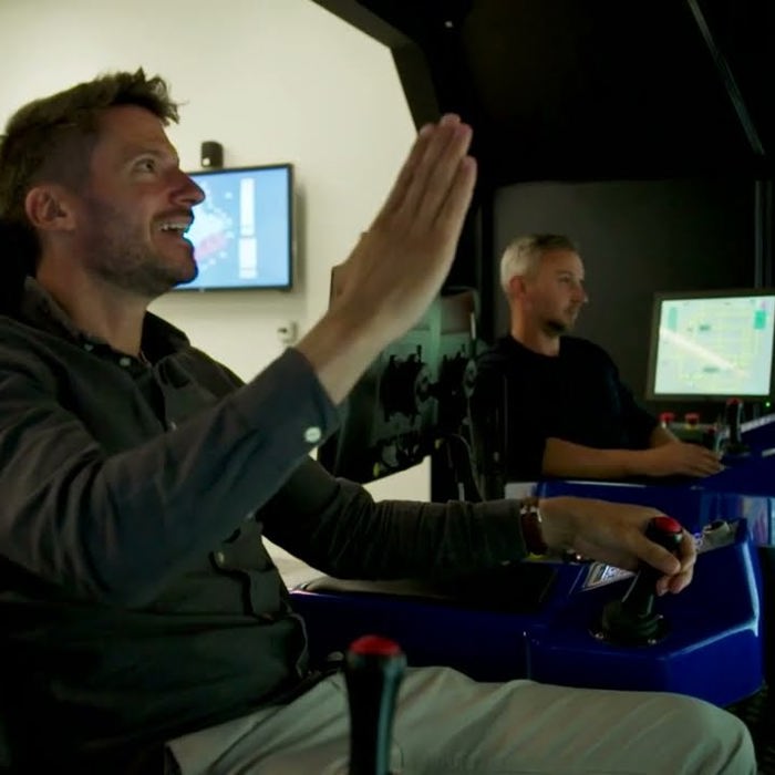 Two men are operating a flight simulator in a control room, with one raising his hand while both focus on screens and controls in front of them.