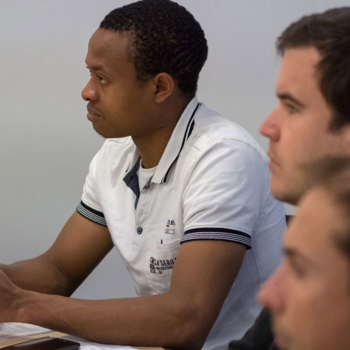 Three men sit at a table, attentively focusing on something off-camera. The man on the left holds a pen and wears a white collared shirt, and they are engaged in a discussion or meeting.