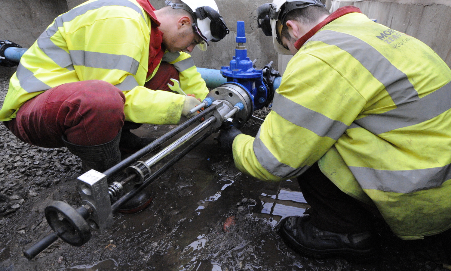 Two workers in high-visibility jackets and helmets, demonstrating their training, repair a pipeline in a muddy outdoor setting.
