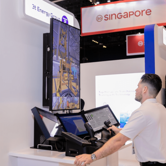A man operates a control system at a trade show booth with a "Singapore" sign overhead and multiple monitor displays. Other attendees and booths are visible in the background.