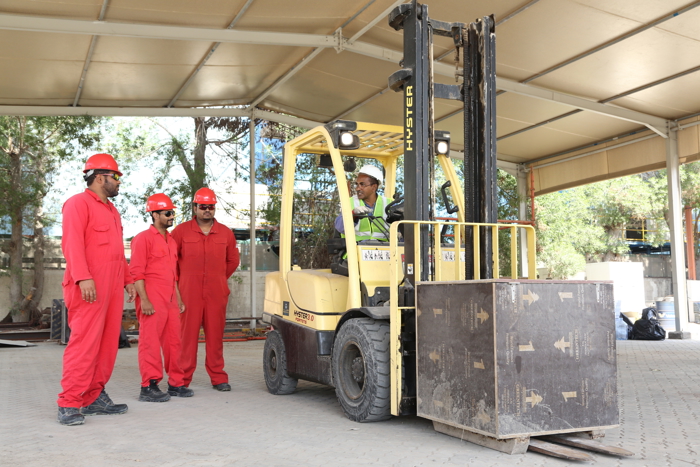 A forklift operator lifts a large box while three workers in red overalls and hard hats watch under a covered area.