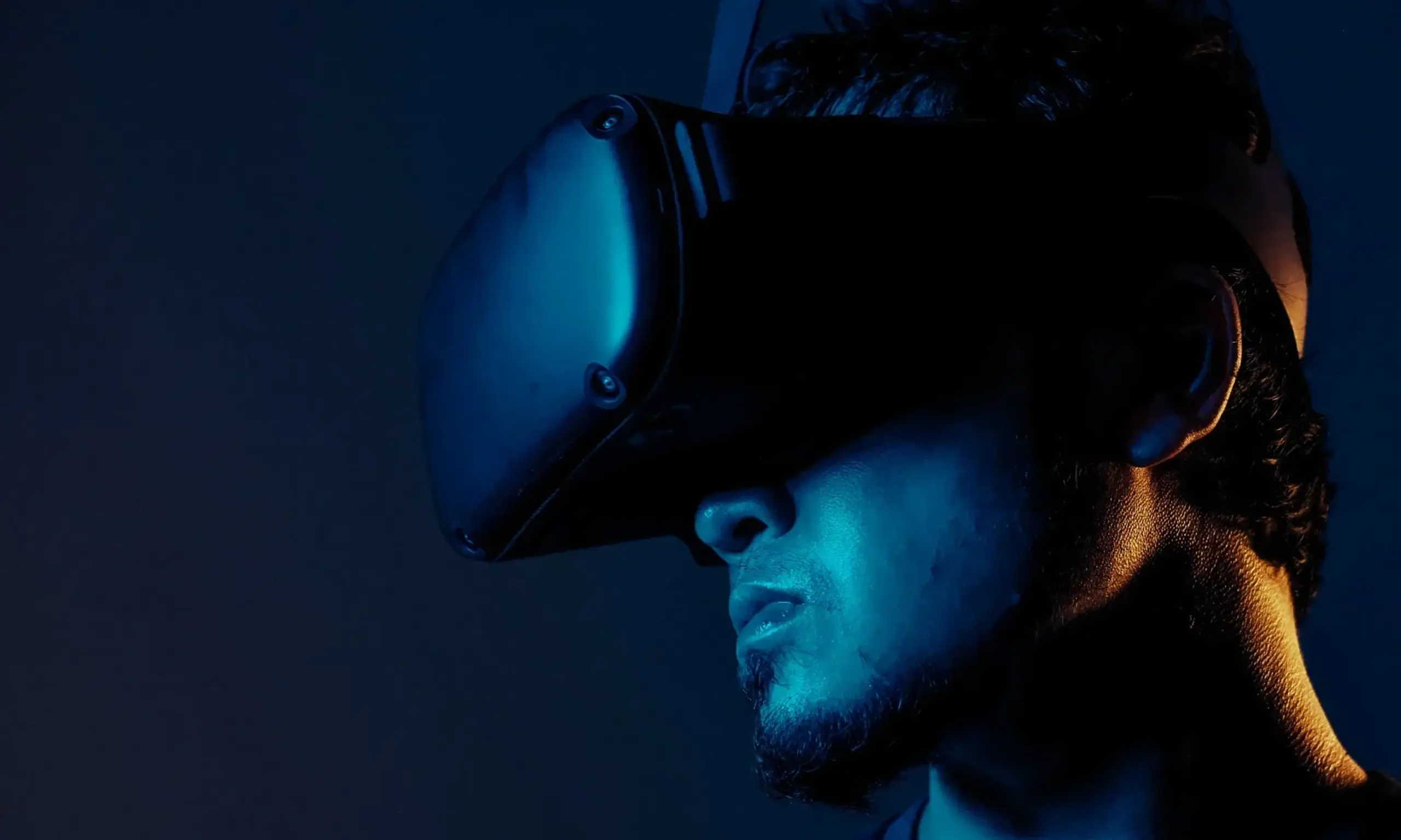 Un hombre con pelo corto y barba que lleva un casco de realidad virtual, iluminado con luz azul sobre un fondo oscuro.