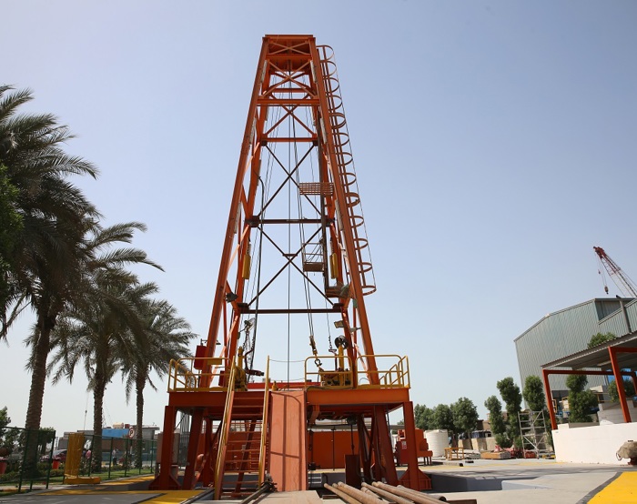 An orange drilling rig stands surrounded by trees with a clear sky in the background. Pipes are visible in the foreground.
