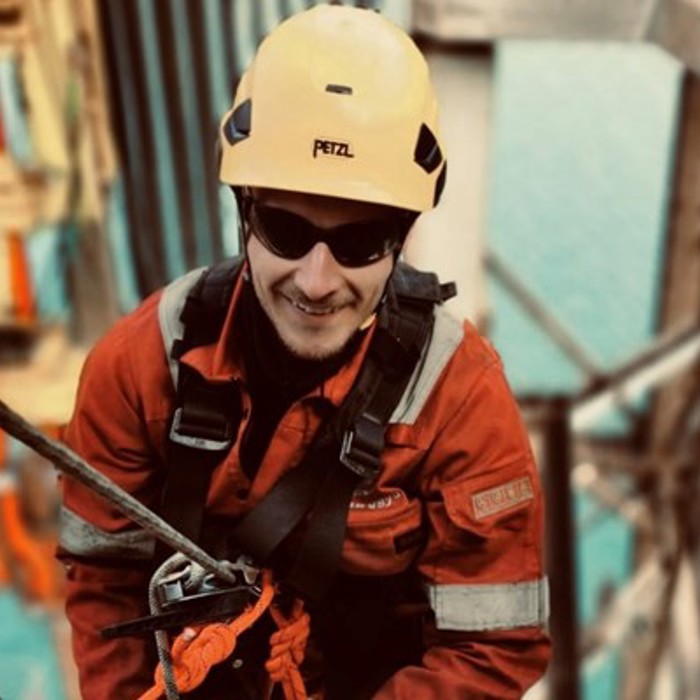 Um trabalhador com um capacete amarelo e equipamento de segurança está a subir uma estrutura com cordas, com uma grande massa de água visível ao fundo.