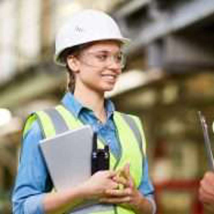 Two workers wearing hard hats and high-visibility vests have a conversation in an industrial setting. One holds a clipboard, and the other holds a wrench.