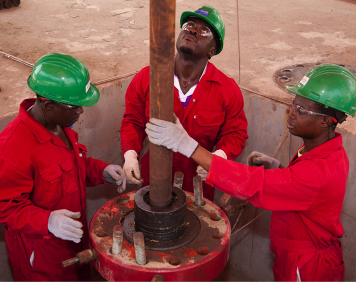 Three workers in red coveralls and green helmets are working with a large pipe in an industrial setting. Two are holding the pipe while one monitors, all wearing safety glasses and gloves.